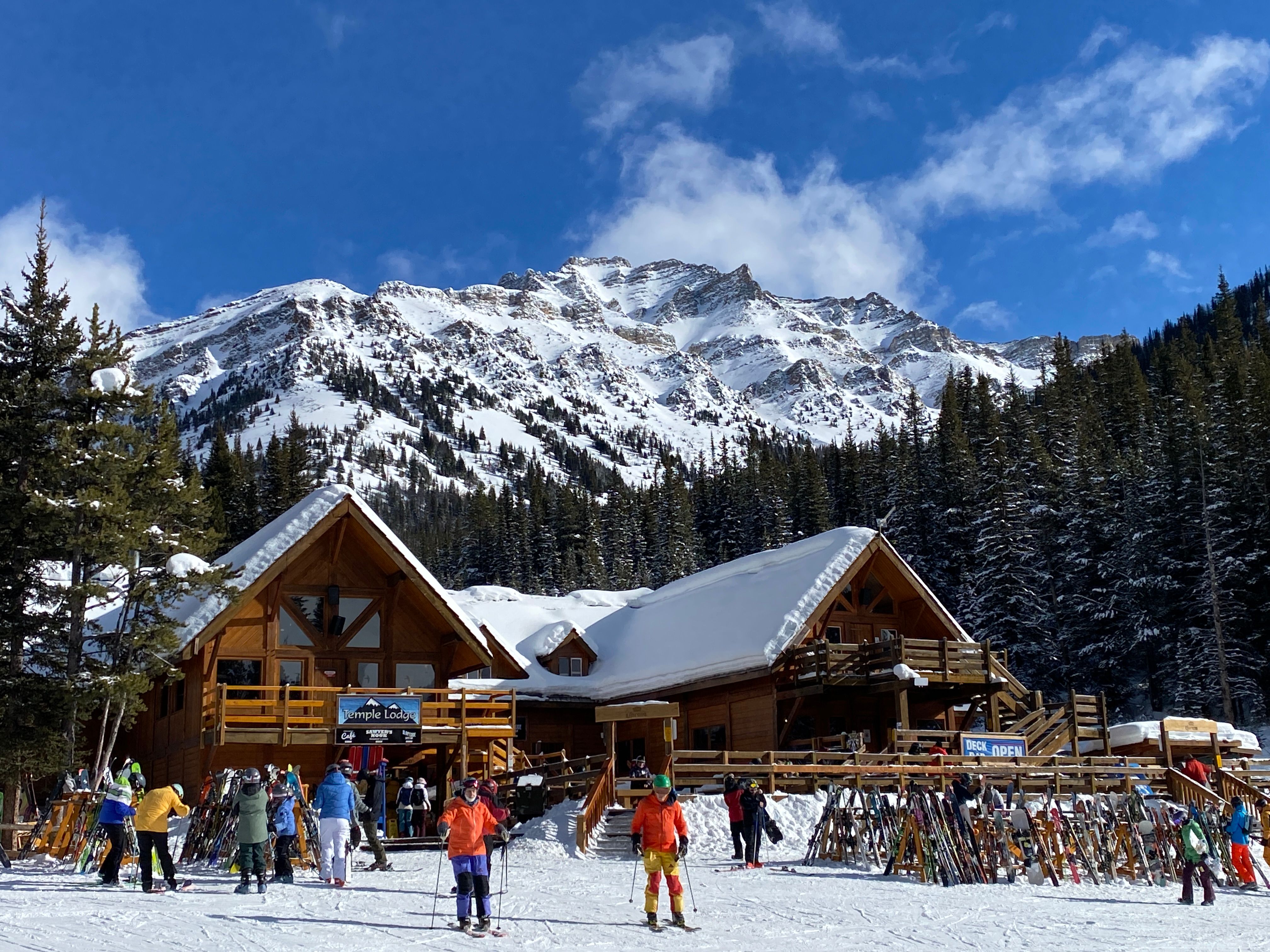 Skiers and snowboarders gather outside a timber lodge with racks of skis, with snow-covered peaks and pine forest behind under a blue sky