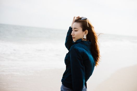 Young woman in a stylish outfit enjoying a breezy day at the beach, with ocean waves in the background.