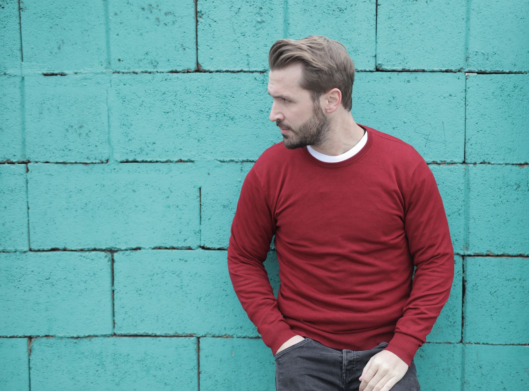 Homme en pull col rond bordeaux adossé à un mur urbain, look streetwear épuré
