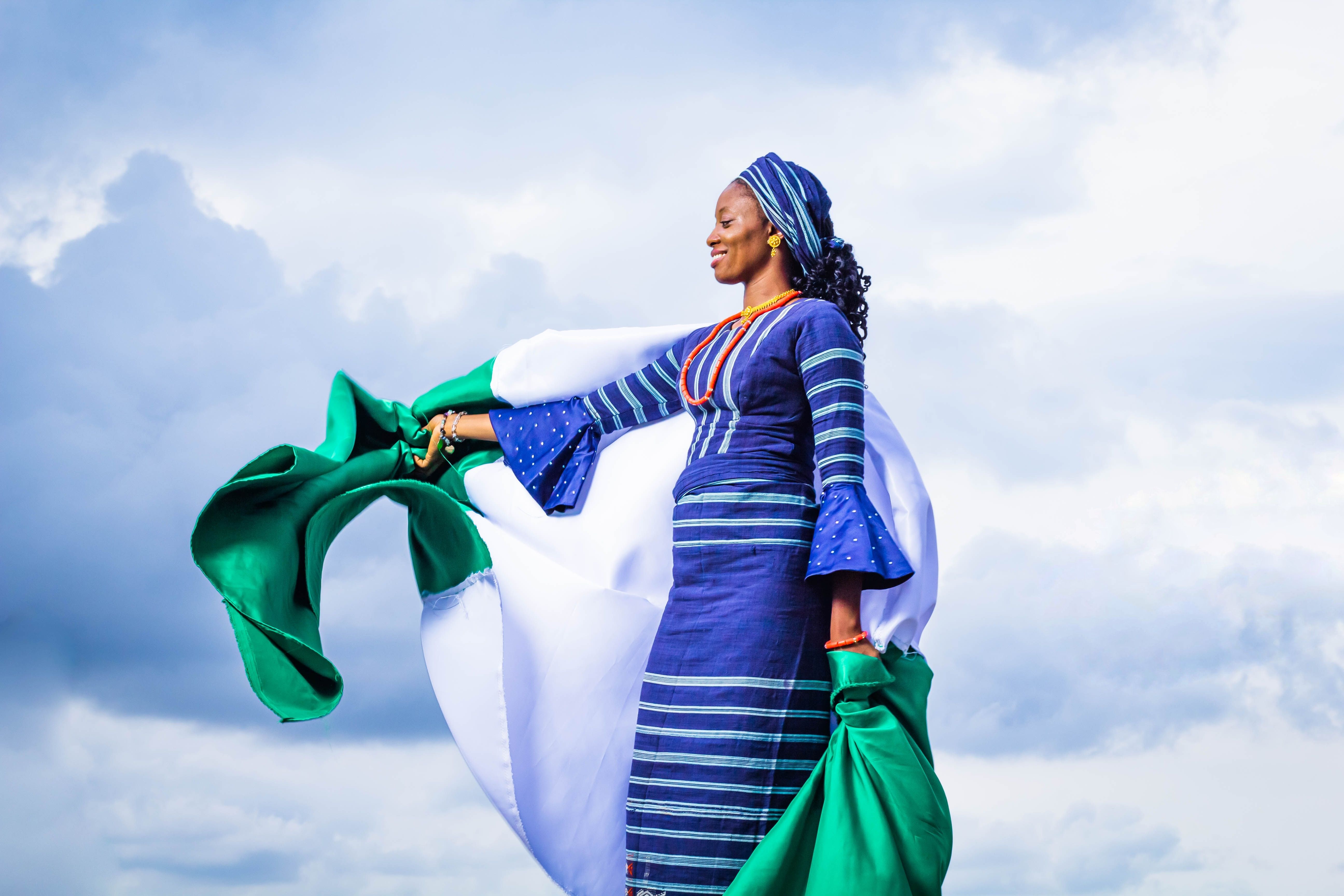 Woman in vibrant traditional wear joyfully holds flag against cloudy sky.
