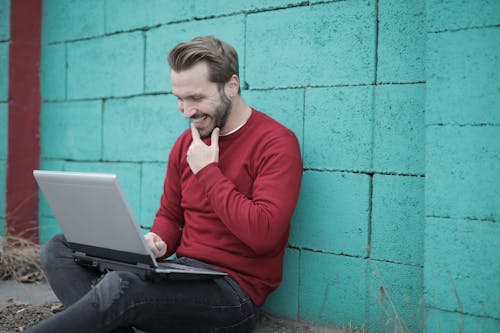 Man in Red Long Sleeve Shirt and Black Denim Jeans Sitting on the Ground Using Laptop