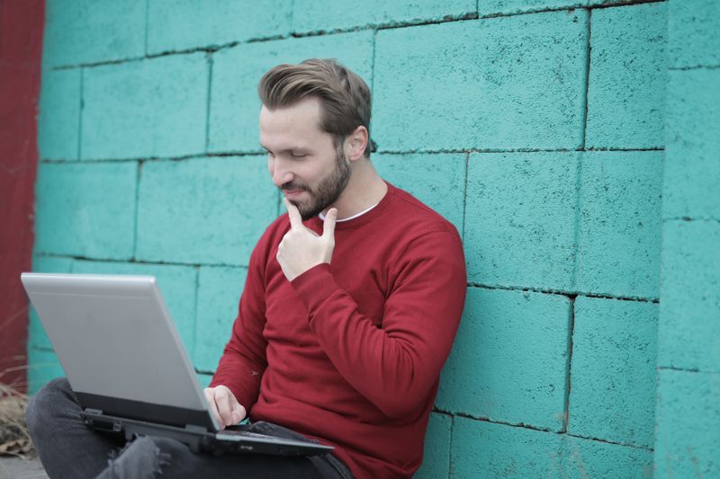 Smiling man using laptop for remote work, leaning on a vibrant green wall outdoors.
