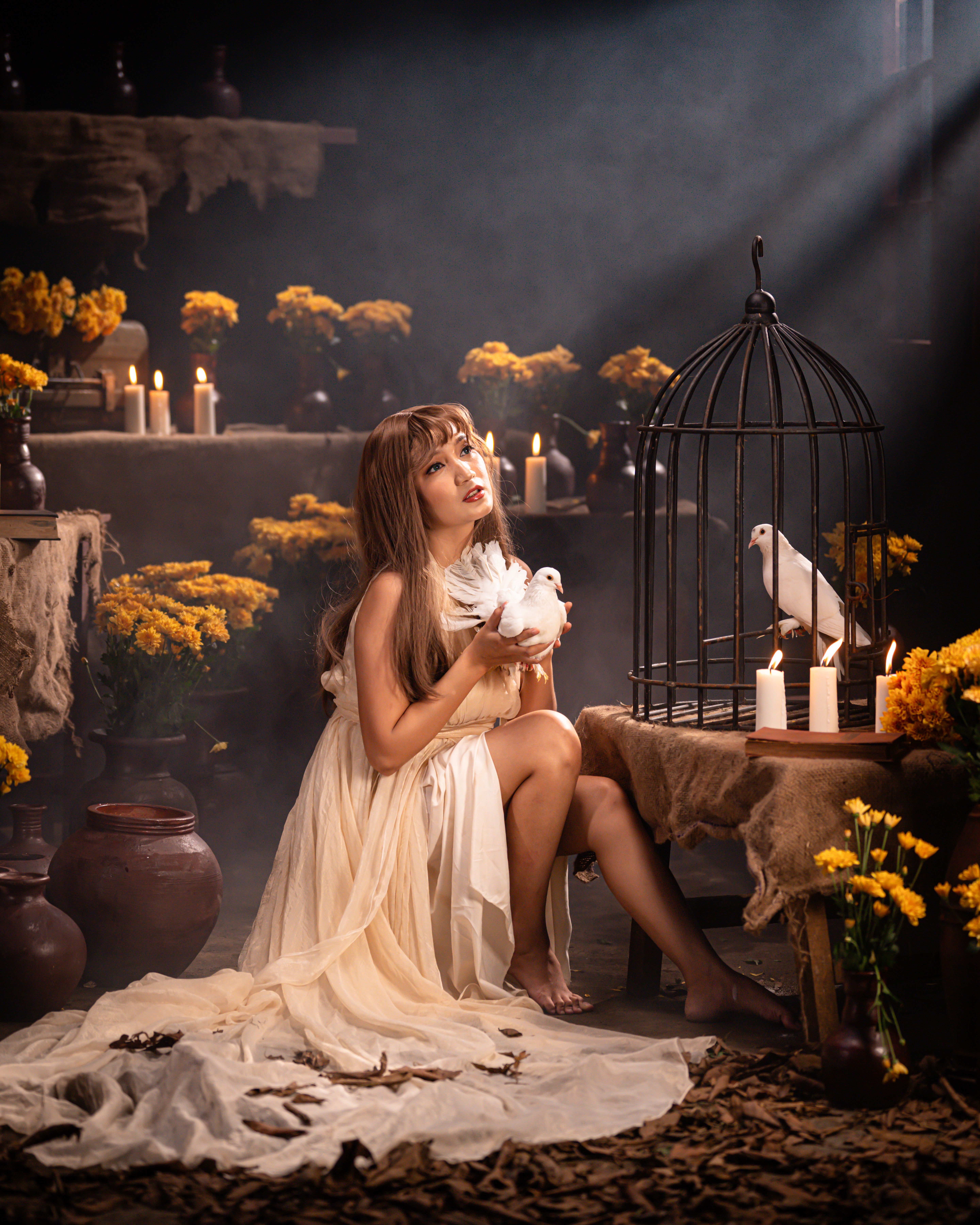 An enchanting woman in a flowing dress holds doves in a candlelit room surrounded by flowers.