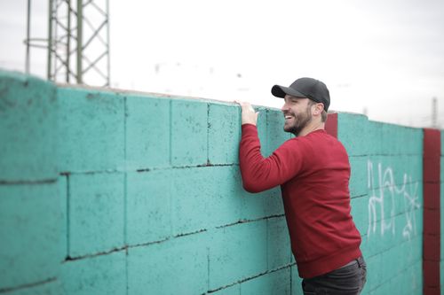 Free Cheerful young bearded man in casual clothes and cap looking over blue brick wall with graffiti on street in city Stock Photo