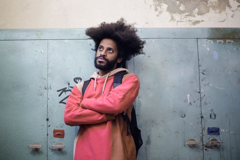 Man with afro hair in a casual hoodie leans pensively on worn lockers indoors. Man with afro hair in a casual hoodie leans pensively on worn lockers indoors.