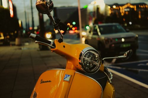 Free Close-up of a yellow Vespa parked on a Warsaw street during twilight with city lights in the background. Stock Photo