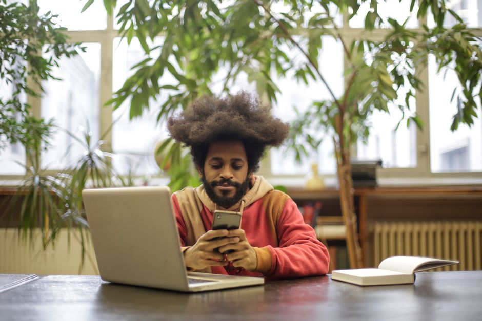 Man with afro-textured hair using laptop and smartphone in a library setting, surrounded by greenery.