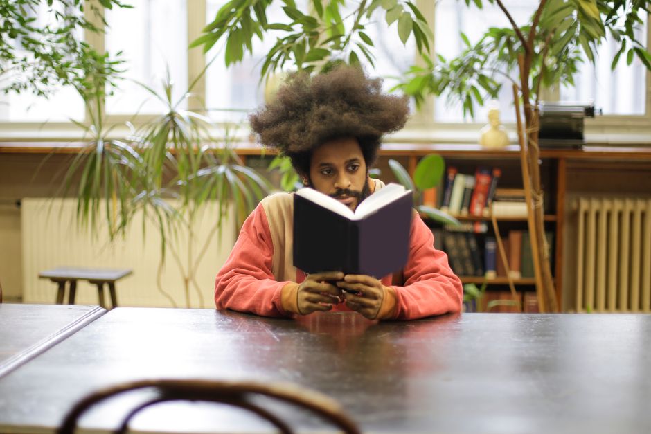 Serious young bearded ethnic man in casual hoodie reading interesting book while sitting at wooden table in modern light library Serious young bearded ethnic man in casual hoodie reading interesting book while sitting at wooden table in modern light library