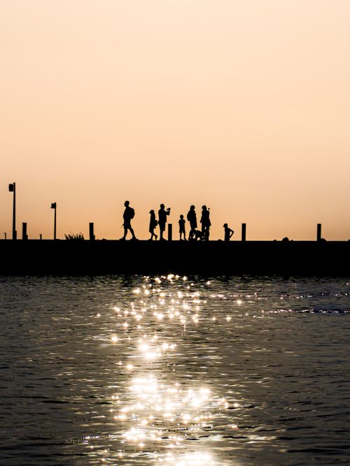 Free A picturesque silhouette of people walking along a pier with sparkling water reflecting the sunset. Stock Photo