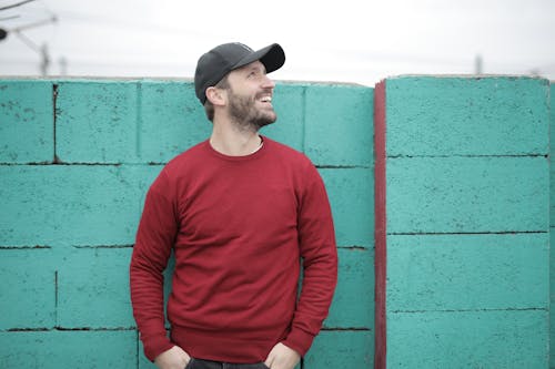 Free Cheerful young bearded man in red sweatshirt and cap standing with hands in pockets near blue brick wall and looking up on street Stock Photo