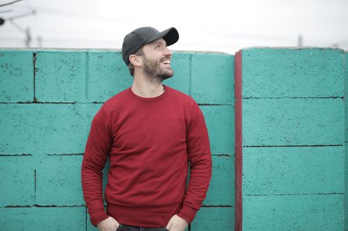 Free Cheerful young bearded man in red sweatshirt and cap standing with hands in pockets near blue brick wall and looking up on street Stock Photo