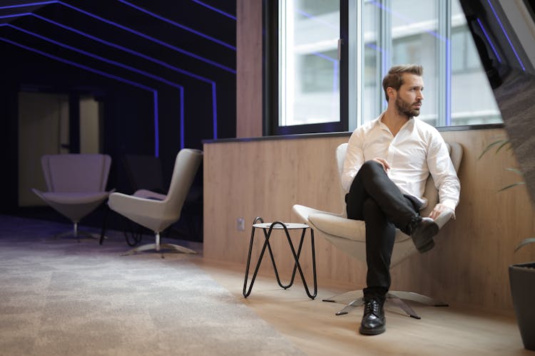 Confident Businessman Sitting On Armchair Near Window In Modern Spacious Workspace