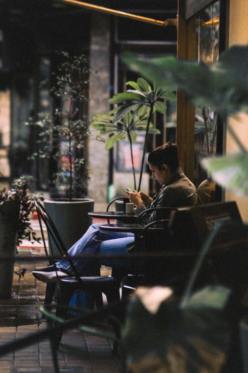 Free A person sitting at a cozy cafe table, engrossed in their smartphone amid lush greenery. Stock Photo