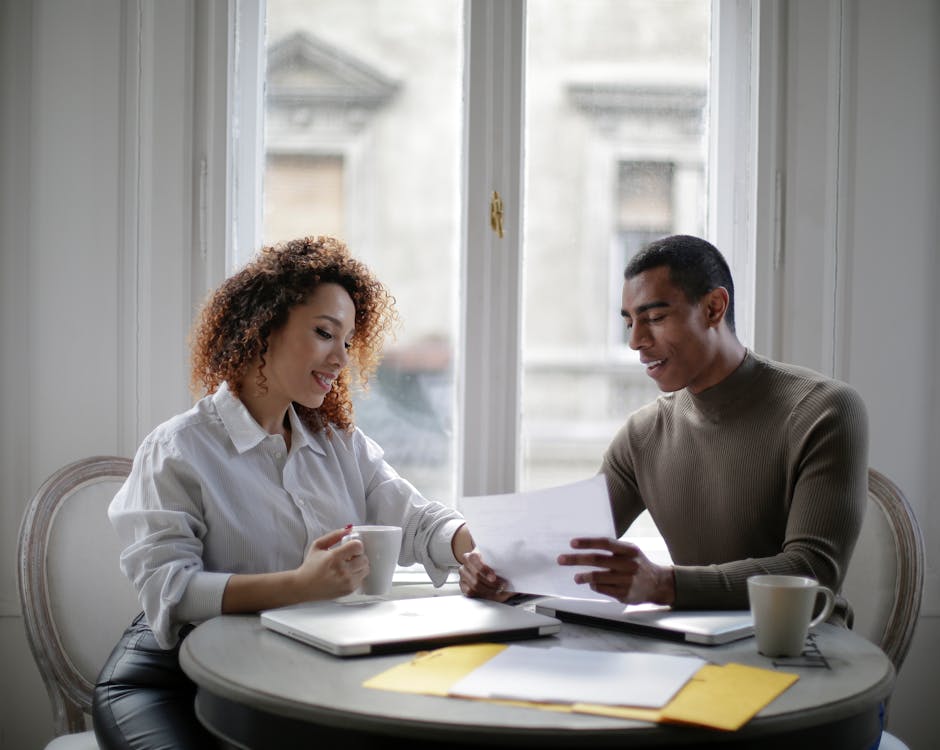 Free Contemporary young diverse couple working together with documents and laptops while sitting at round table near window and drinking coffee from white cups at home Stock Photo
