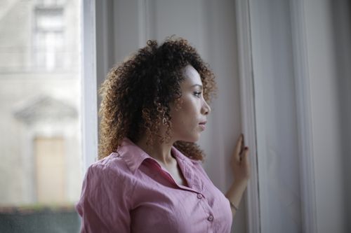 Free Pensive young ethnic lady in casual pink shirt standing near window and looking away thoughtfully while resting in modern light apartment Stock Photo