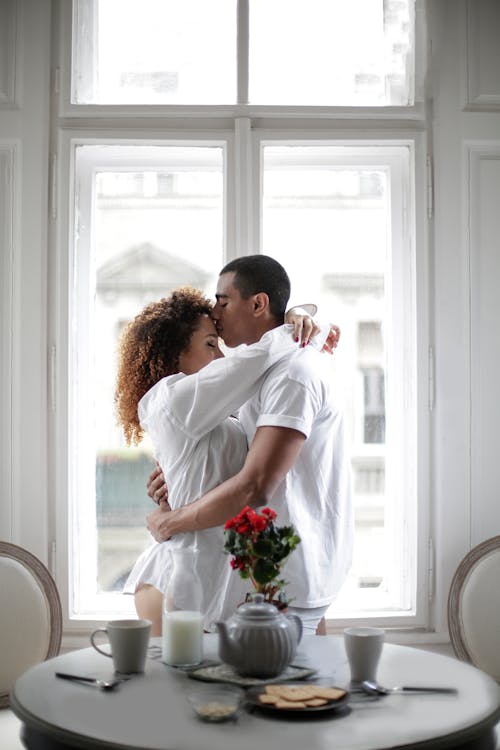 Free Couple Hugging And Kissing While Standing Near Window At Home Stock Photo