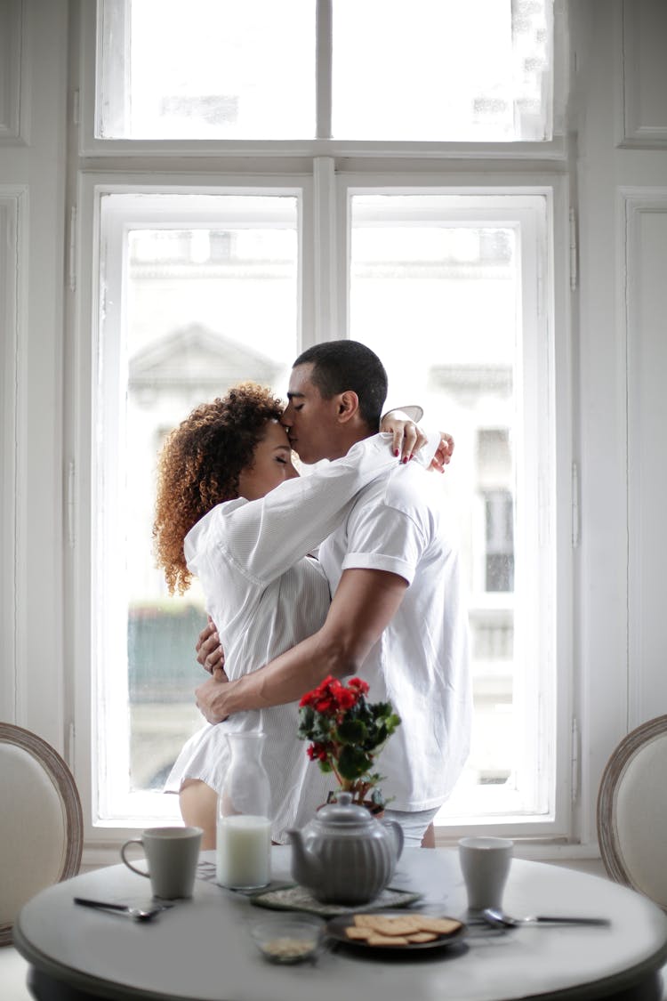 Couple Hugging And Kissing While Standing Near Window At Home