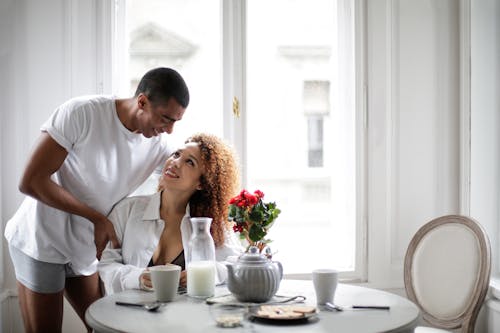 Free Smiling young ethnic couple in underwear clothes gathering around table near window while having breakfast and looking at each other at light modern apartment Stock Photo