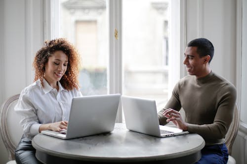 Cheerful young couple using laptops while sitting at table near window at home