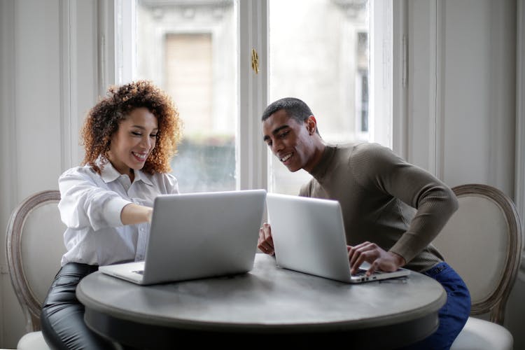 Positive Young Ethnic Colleagues Using Laptops On Round Table Near Window At Home