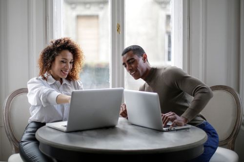 Free Smiling young ethnic couple wearing casual clothes sitting at round table near window and working on laptops while discussing details of project at home Stock Photo