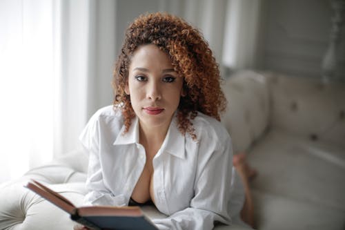 Free Sensual ethnic female in white shirt with book resting on comfortable sofa in light living room and looking at camera Stock Photo