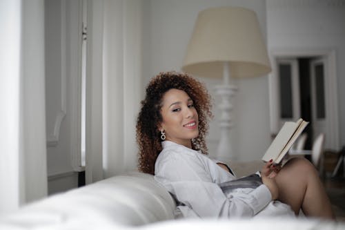 Woman in White Long Sleeve Shirt Sitting on Couch Holding a Book