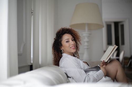 Free A woman with curly hair reads a book on a comfy sofa in a bright, stylish living room. Stock Photo