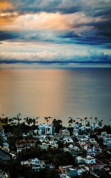 Aerial view of a tranquil coastal village with palm trees and stunning ocean scenery under dramatic clouds.