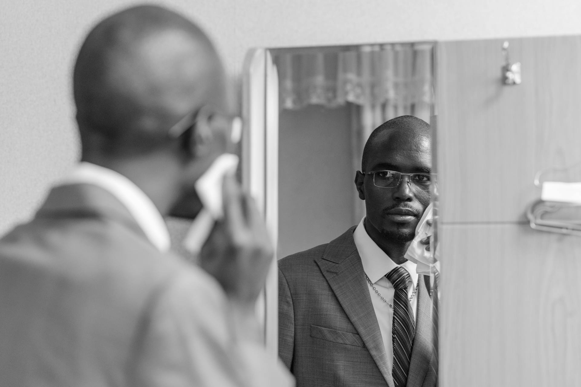 Black and white portrait of a man in a suit adjusting his tie in the mirror, creating a sophisticated reflection.