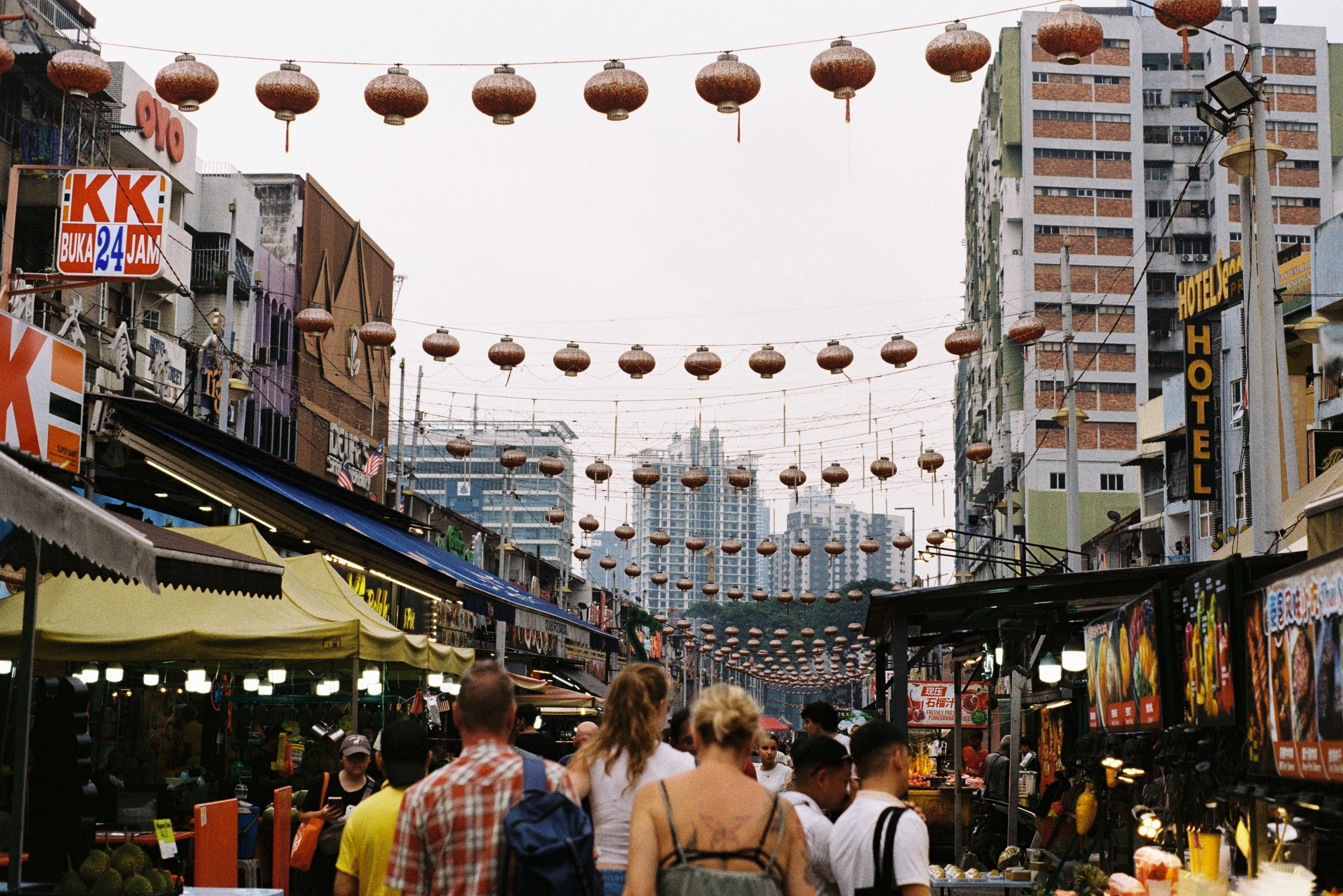 A crowded street of food stalls under strings of paper lanterns, between tall buildings