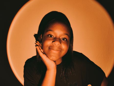 A smiling girl posing with her hand on cheek against a warm orange background.
