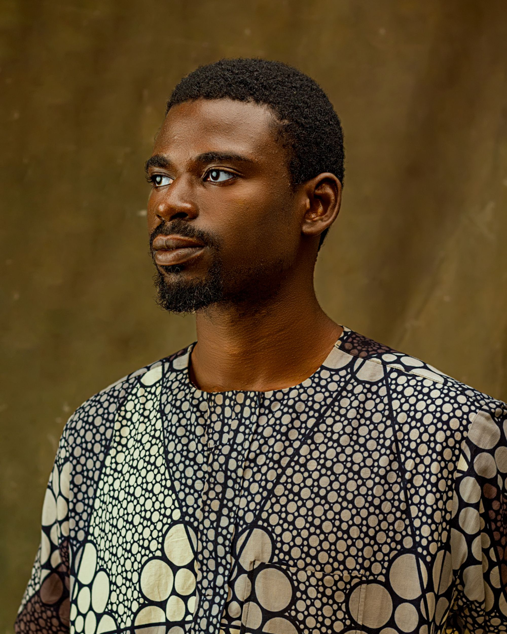A thoughtful adult man gazing sideways, wearing a patterned traditional shirt against a plain background.