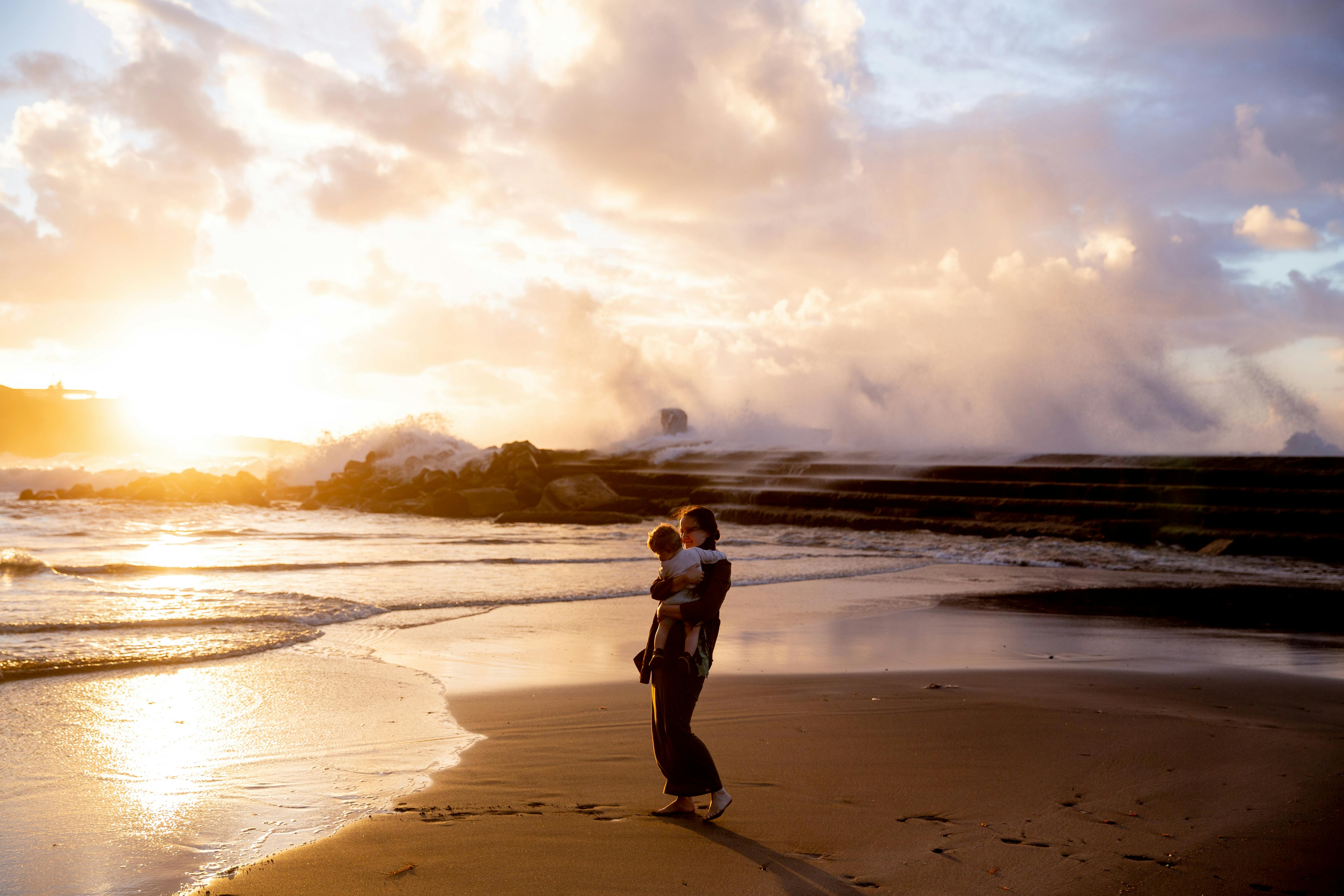 woman standing on seashore carrying her child during sunset