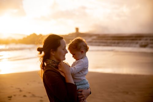 Free A mother lovingly holds her baby at a scenic beach during sunset, capturing affection and warmth. Stock Photo