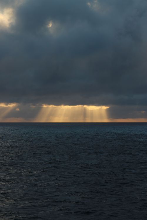 Free Stunning ocean view with sun rays piercing through dramatic clouds during sunset. Stock Photo