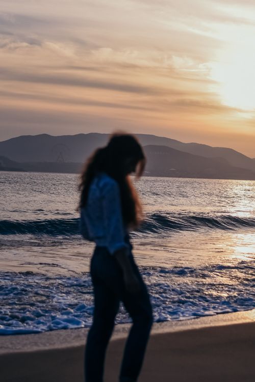 Free A woman walks along the beach at sunset with waves gently crashing and mountains in the background. Stock Photo