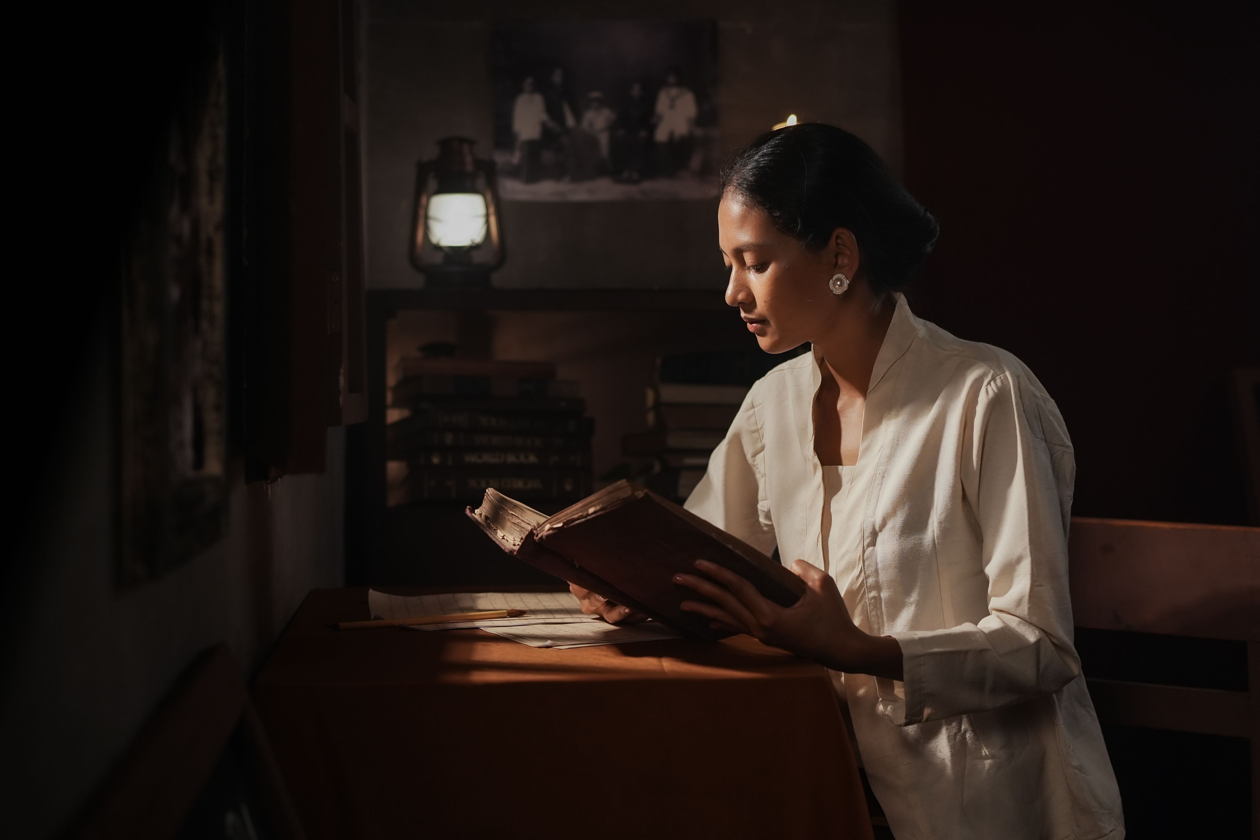 A woman in traditional attire reads an old book indoors by lantern light.