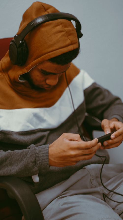 Free A teenager in a hoodie and headphones concentrates while listening to music on a smartphone indoors. Stock Photo