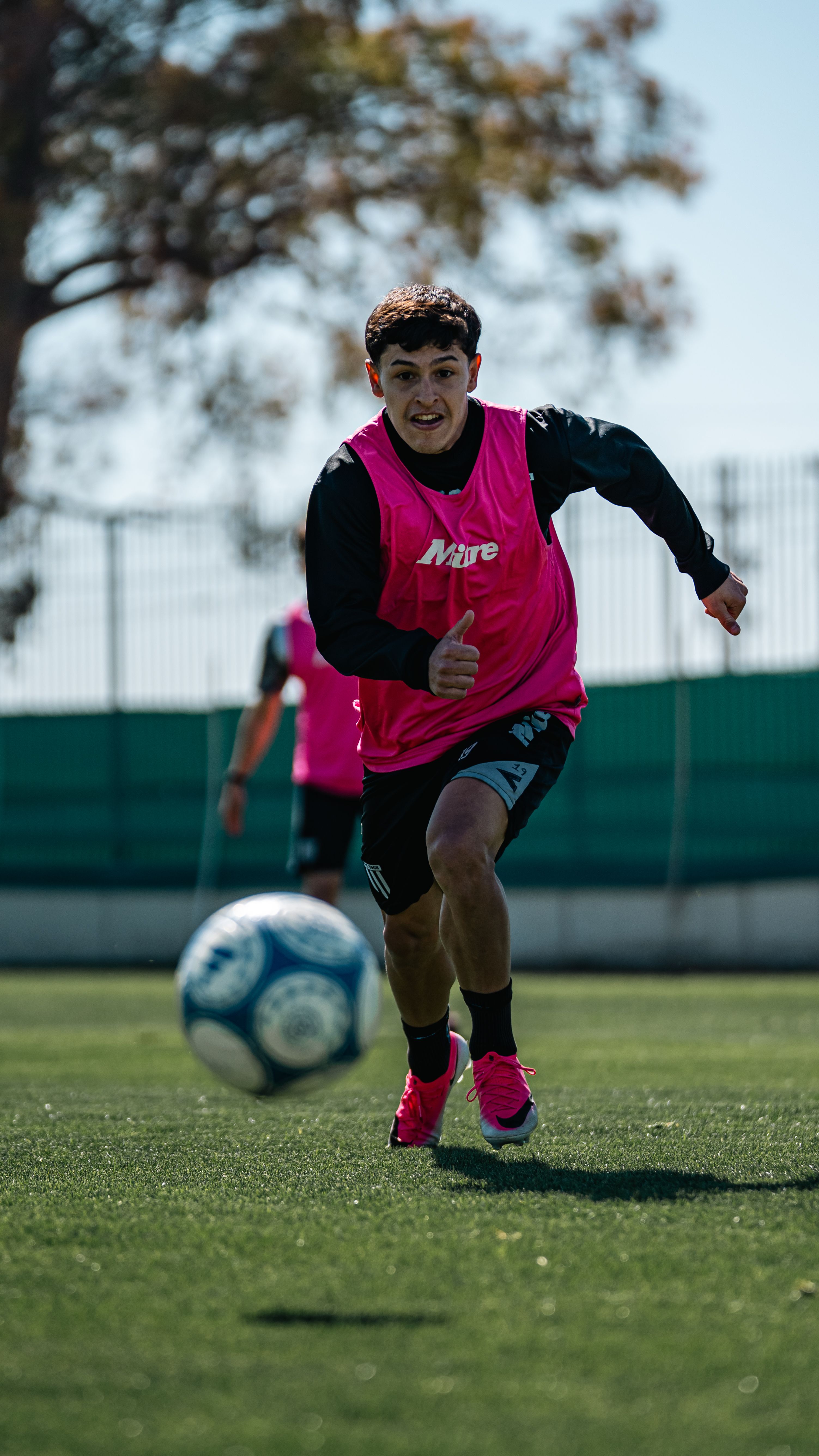 Male soccer player in action on the field wearing a pink vest and sprinting towards the ball.