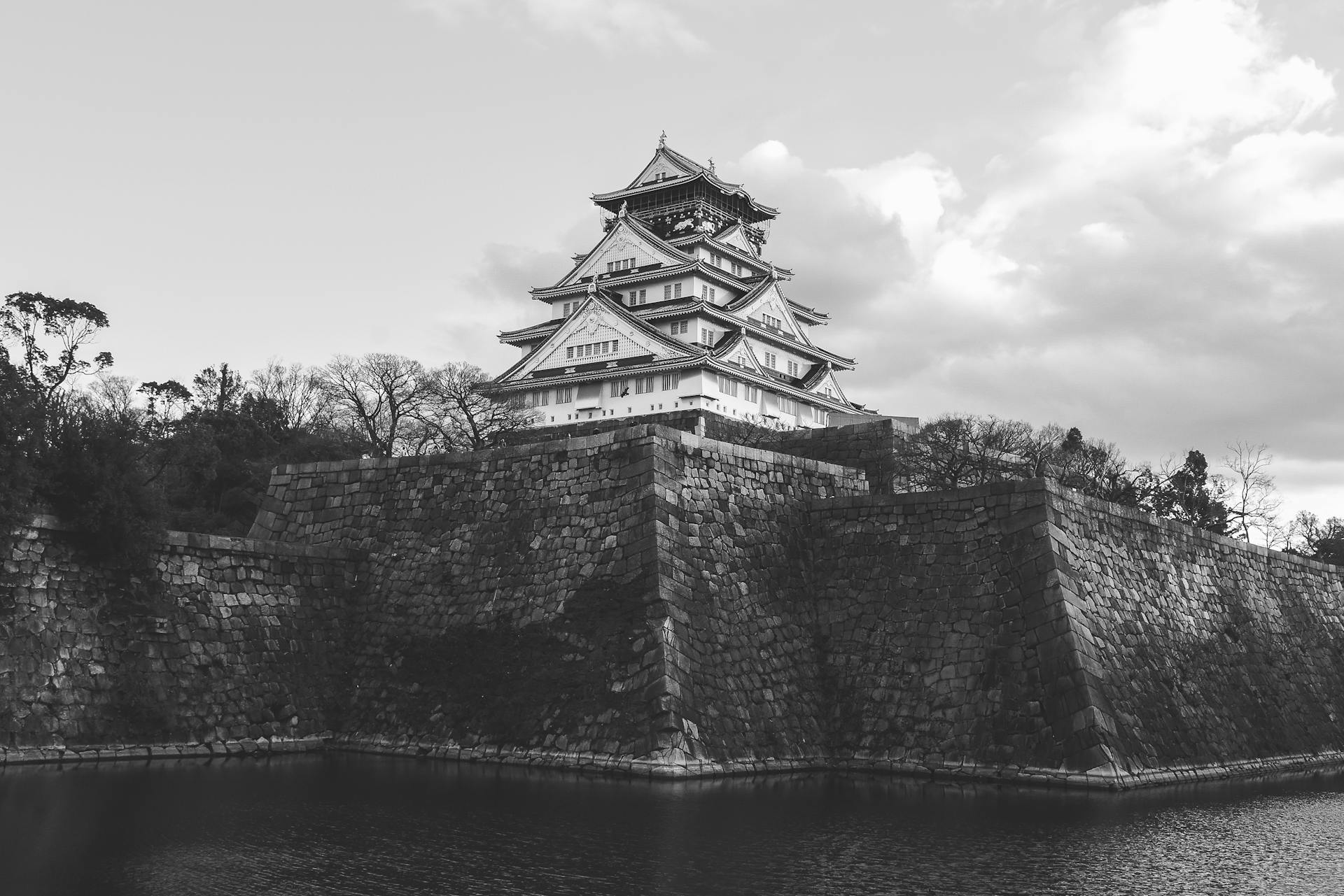 A dramatic black and white image of Osaka Castle, showcasing traditional Japanese architecture against a cloudy sky.