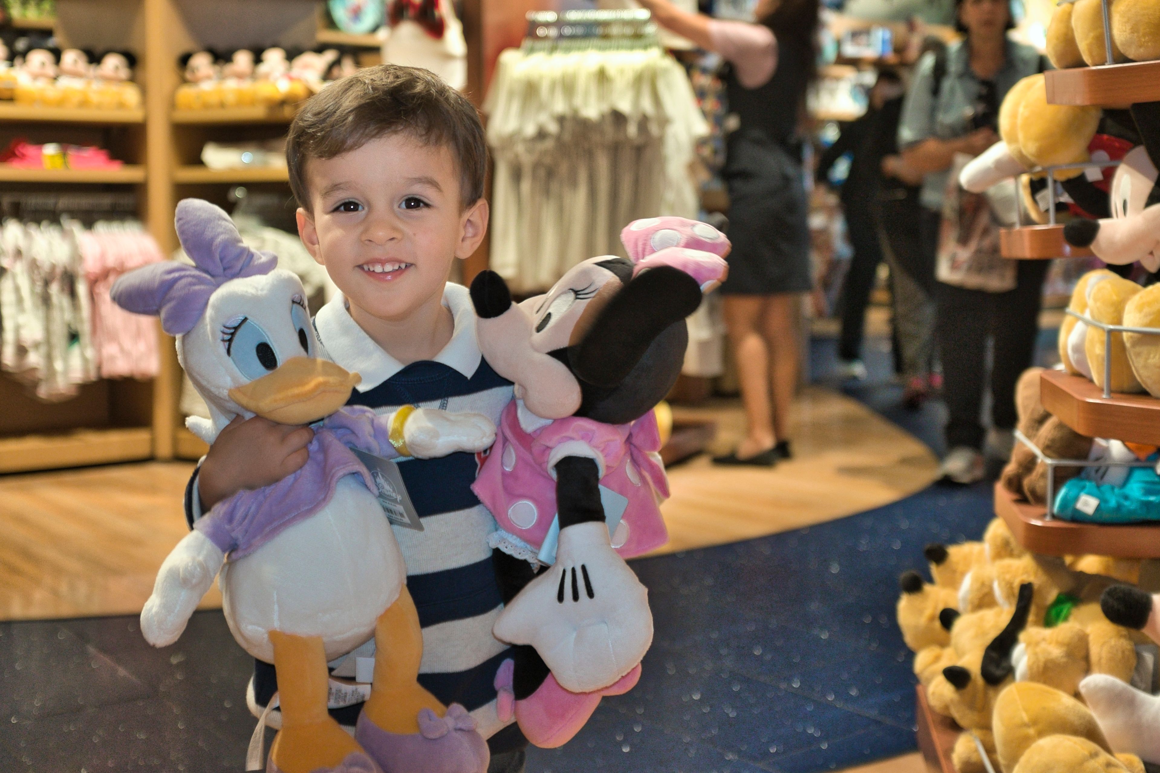 Young boy happily holding Minnie Mouse and Daisy Duck plush toys in a Disney-themed store.