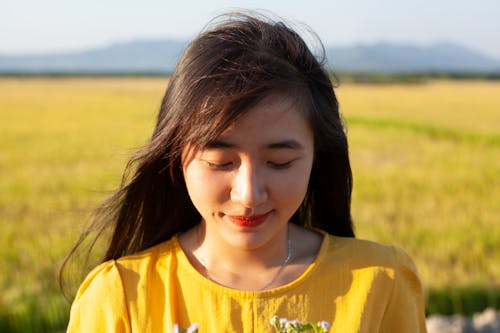Positive young Asian female with long dark hair in summer dress relaxing on grassy field in countryside with closed eyes on sunny day