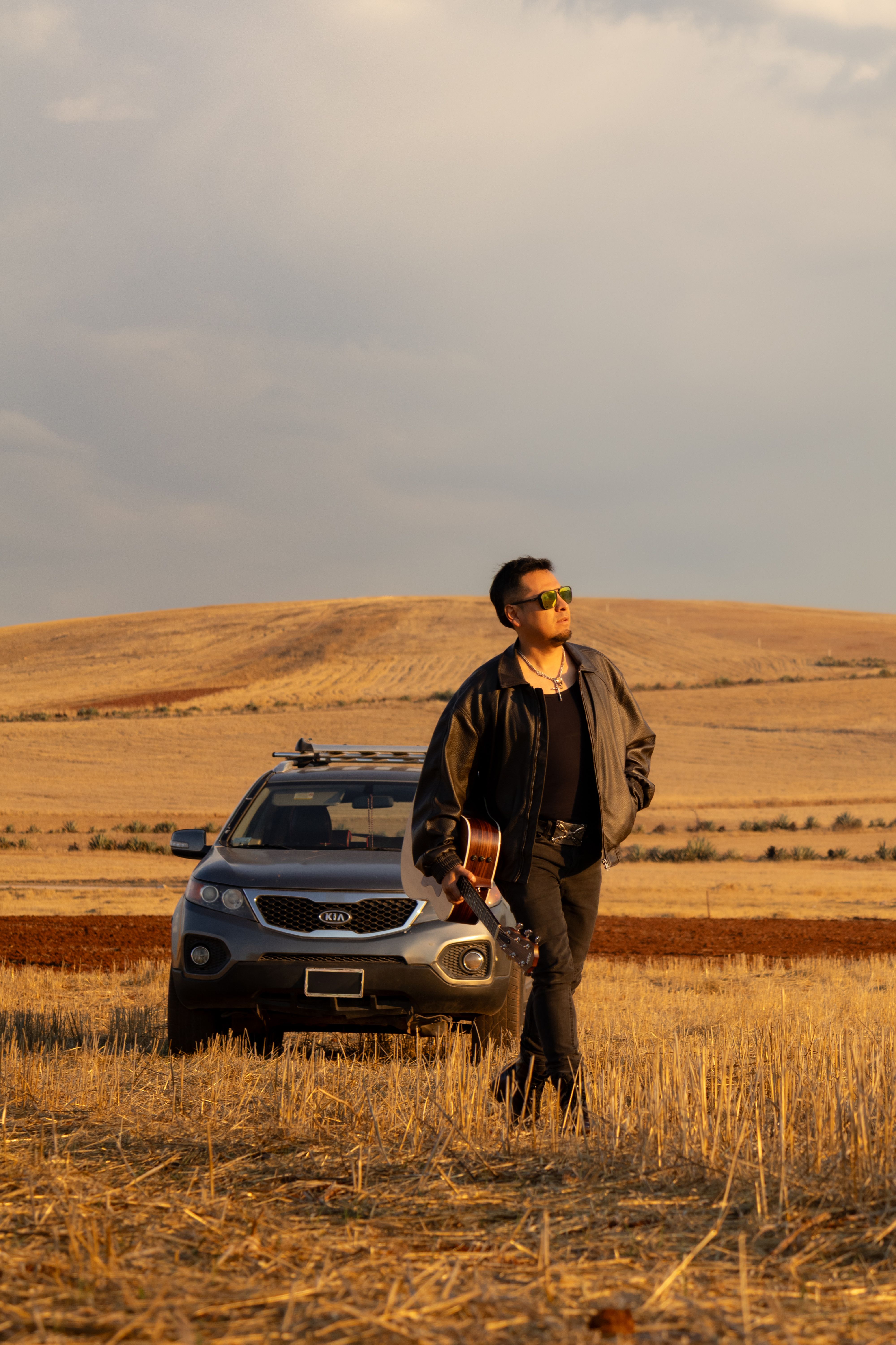 A man with a guitar and sunglasses stands in a field beside a car under a dramatic sky.
