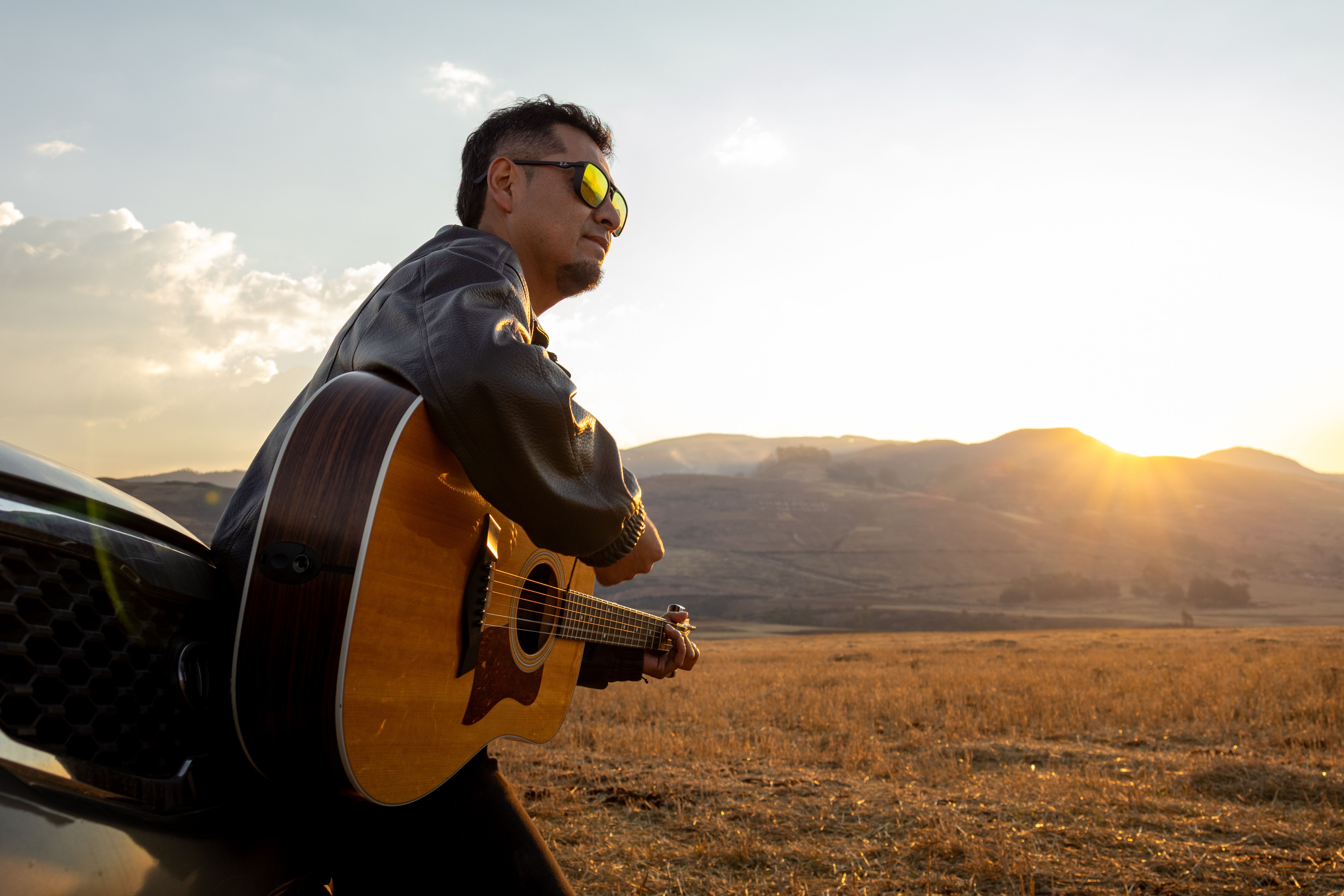 A man leaning on a car, playing a guitar during sunset in a wide open field.