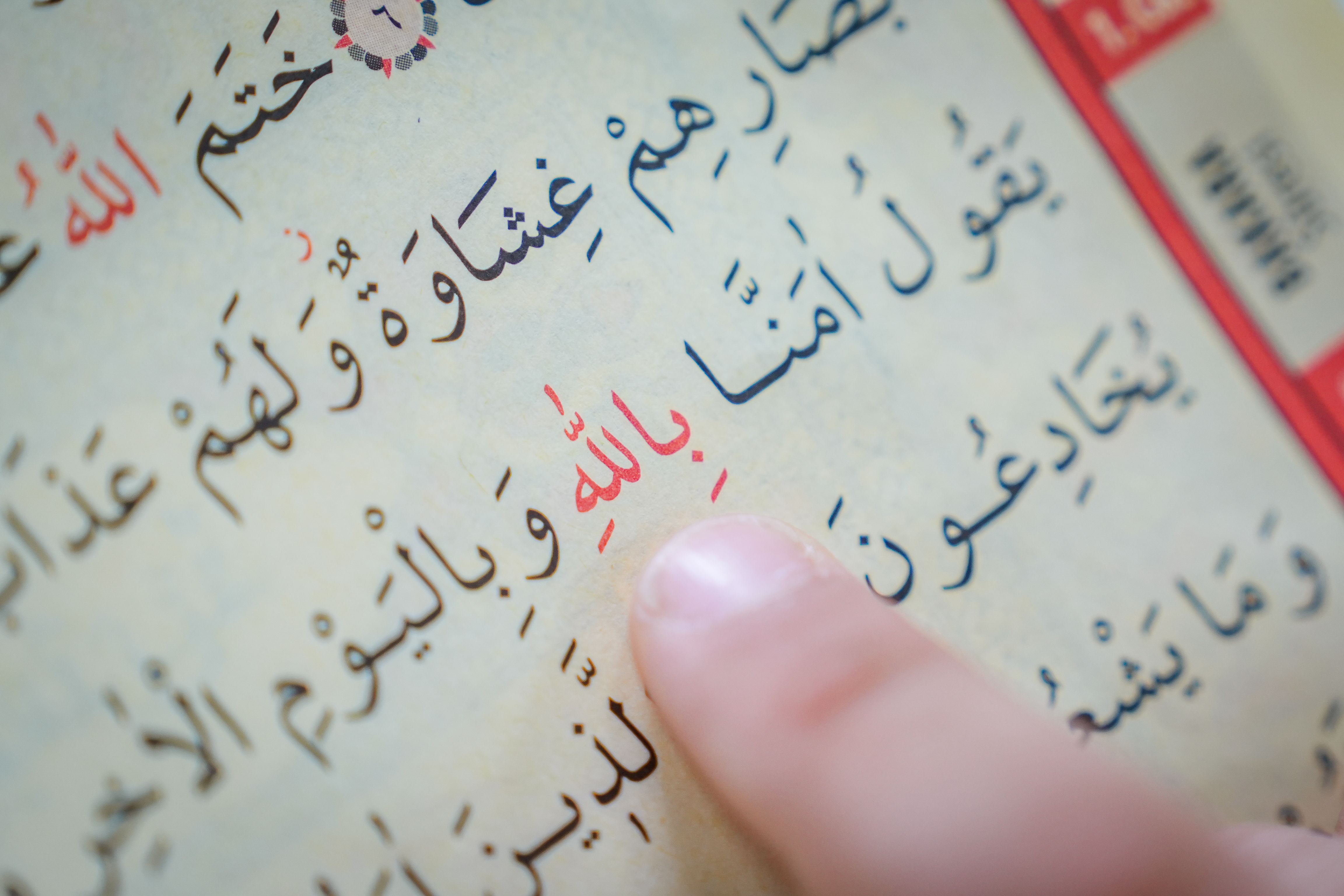 A person's finger pointing at Arabic text in a religious book, illustrating learning or teaching context.