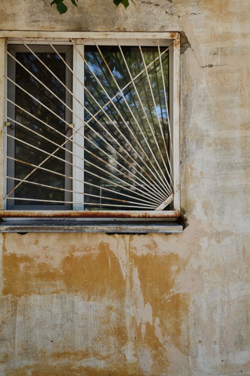 Free A weathered wall with a barred window reflecting trees, creating a rustic aesthetic. Stock Photo