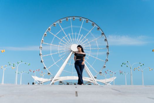 A young woman poses stylishly in front of the Baku Ferris Wheel under a clear blue sky.