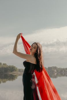 Woman in black dress and red scarf posing by a calm lake.