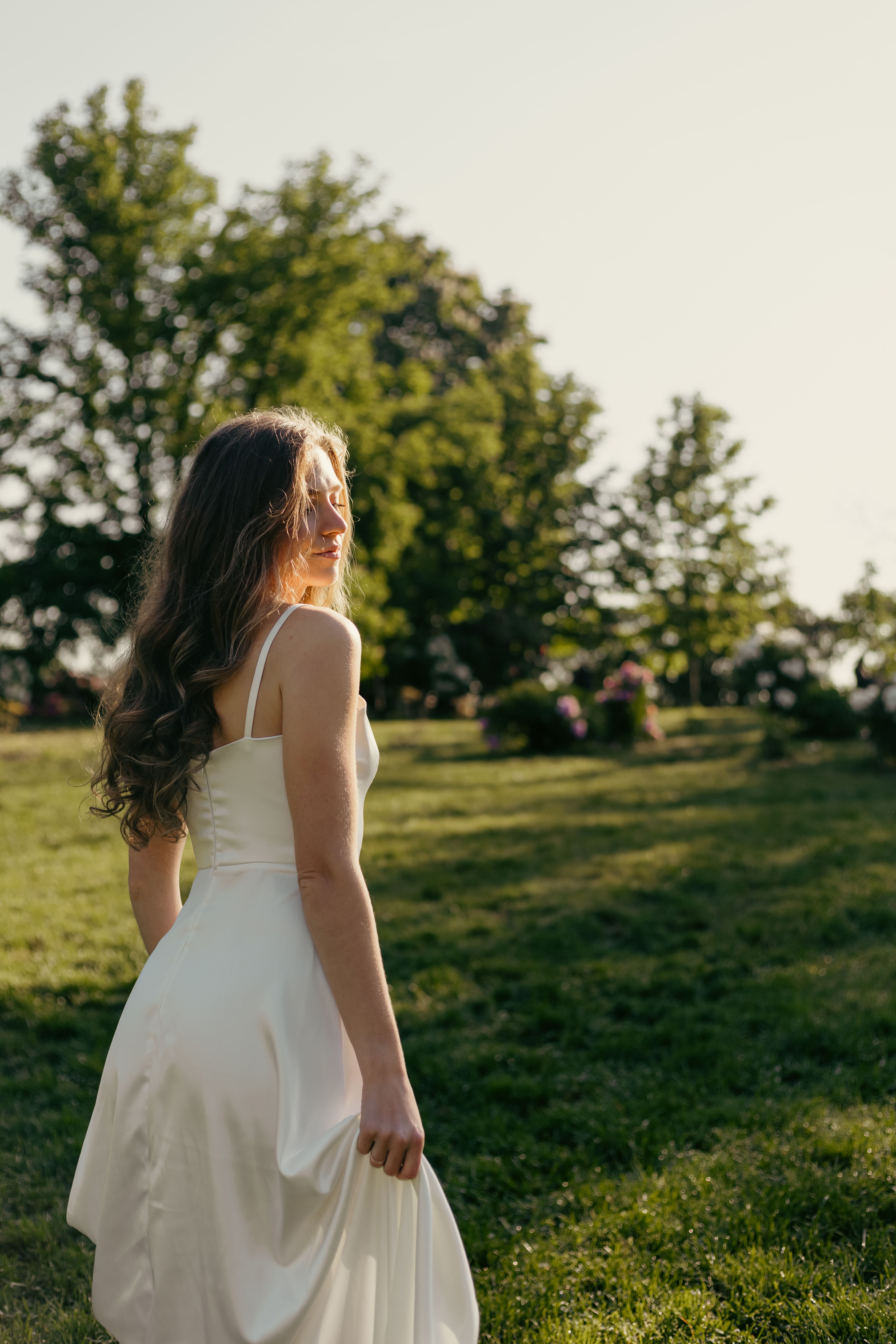 A woman in a white dress enjoys the warm sunlight in a serene park setting.